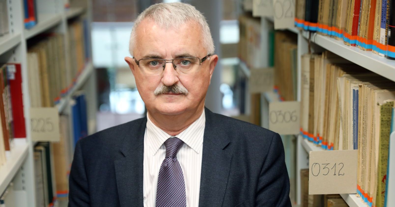 Prof. dr hab. Bogusław Dybaś Potret mężczyzny stojącego między dwoma regałami z książkami w bibliotece.