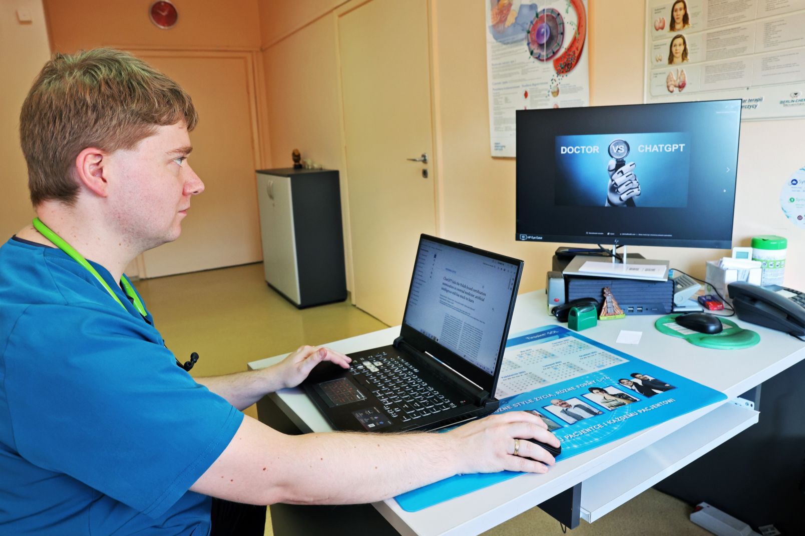 Dr Szymon Suwała siedzi przy laptopie, w tle stoi monitor na którym wyświetla się napis: doctor vs chatgpt