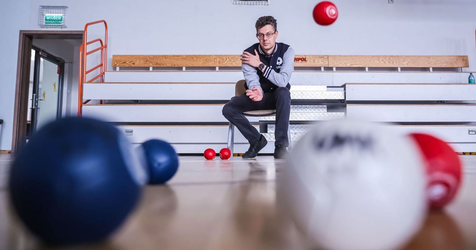 Mikołaj Piekut, coach and specialist in the Support Team for People with Special Needs at the NCU's Centre for Support and Personal Development Mikołaj Piekut sitting on a bench in a gym with scattered boccia balls.