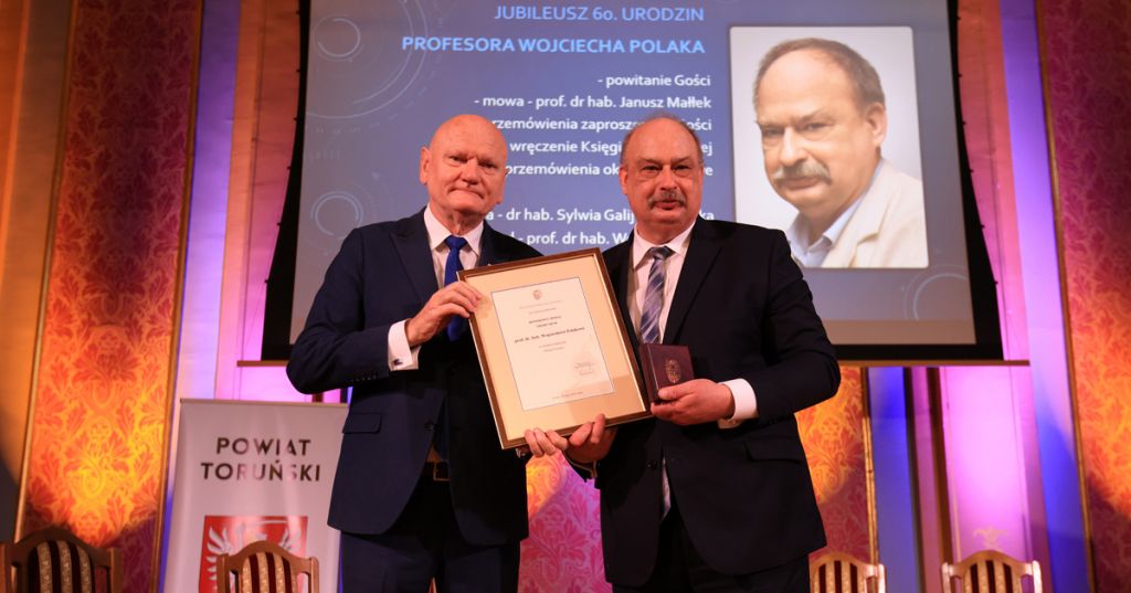 Medal Honorowy "Thorunium", który otrzymał prof. Wojciech Polak, to najwyższe wyróżnienie prezydenckie Prezydent Zaleski i prof. Polak pozują do zdjęcia po wręczeniu medalu. Prof. Polak w lewej dłoni trzyma medal, a prawą podtrzymuje dyplom w ramce. W tle widać wyświetlony program wydarzenia
