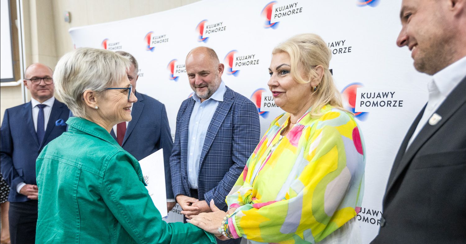 W imieniu Uniwersyteckiego Liceum Ogólnokształcącego potwierdzenie zawarcia umowy odebrała dyrektor dr Beata Trapnell Dr Beata Trapnell i Elżbieta Piniewska podają sobie ręce, obok stoi marszałek Całbecki i inni urzędnicy. W tle widać także materiałową ściankę z logo województwa.