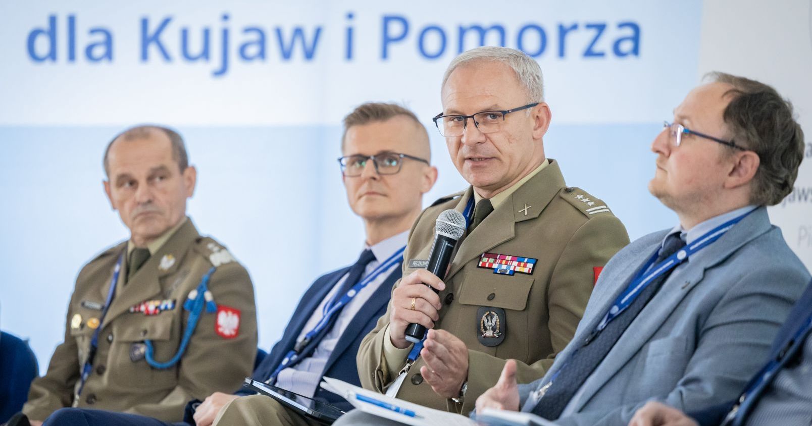 Pomysłodawcą I Forum Bezpieczeństwa i Przemysłu Obronnego był zarząd Fundacji Infosec: ppłk. rez. Jarosław Wiśnicki oraz dr hab. Arkadiusz Czwołek, prof. UMK Zdjęcie czterech mężczyzn podczas panelu dyskusyjnego. Dwóch ma na sobie mundury wojskowe, a pozostali garnitury. Jeden z umundurowanych mężczyzn trzyma mikrofon i przemawia. W tle fragment baneru z napisem "dla Kujaw i Pomorza"