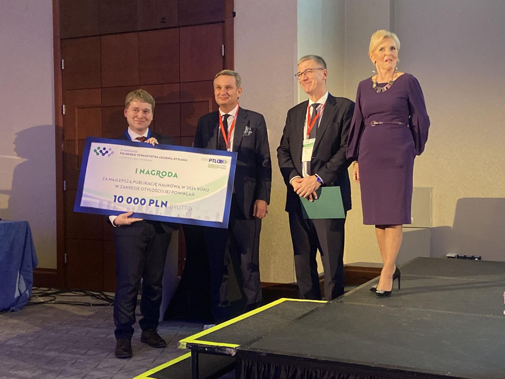 Zdjęcie z uroczystości wręczenia nagr&oacute;d, dr Szymon Suwała stoi z dużym kartonowym czekiem na 10 tys. zł