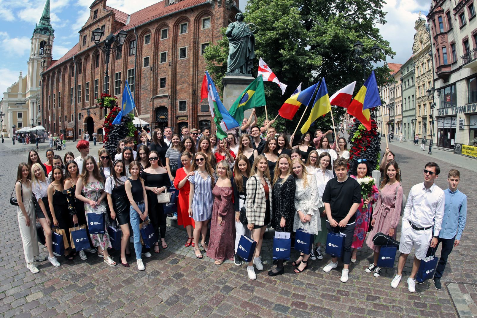 Na zdjęciu stoi grupa ludzi, niektórzy trzymają flagi. Za nimi stoi pomnik Mikołaja Kopernika i Ratusz Staromiejski w Toruniu.