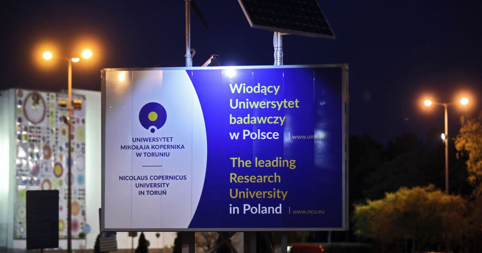  Tablica informacyjna z logo Uniwersytetu i napisem "Wiodący Uniwersytet badawczy w Polsce", w tle widać Aulę UMK