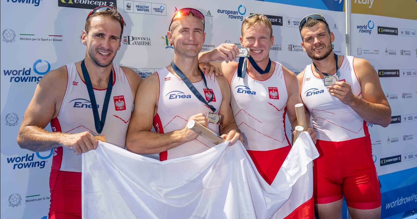 Czterech wioślarzy w strojach narodowych trzyma biało-czerwona flagę. Na szyi mają srebrne medale