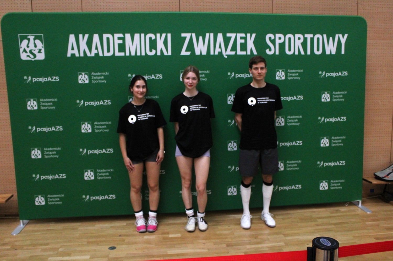 Troje zawodnik&oacute;w UMK stoi na tle zielonej ścianki AZS