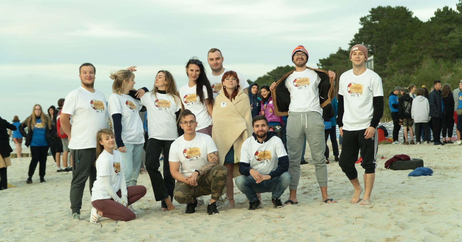 Adapciak to przestrzeń do integracji i tworzenia wspólnych wspomnień z nowo poznanymi ludźmi Grupa osób na plaży w koszulkach z grafiką Adapciaka 2022