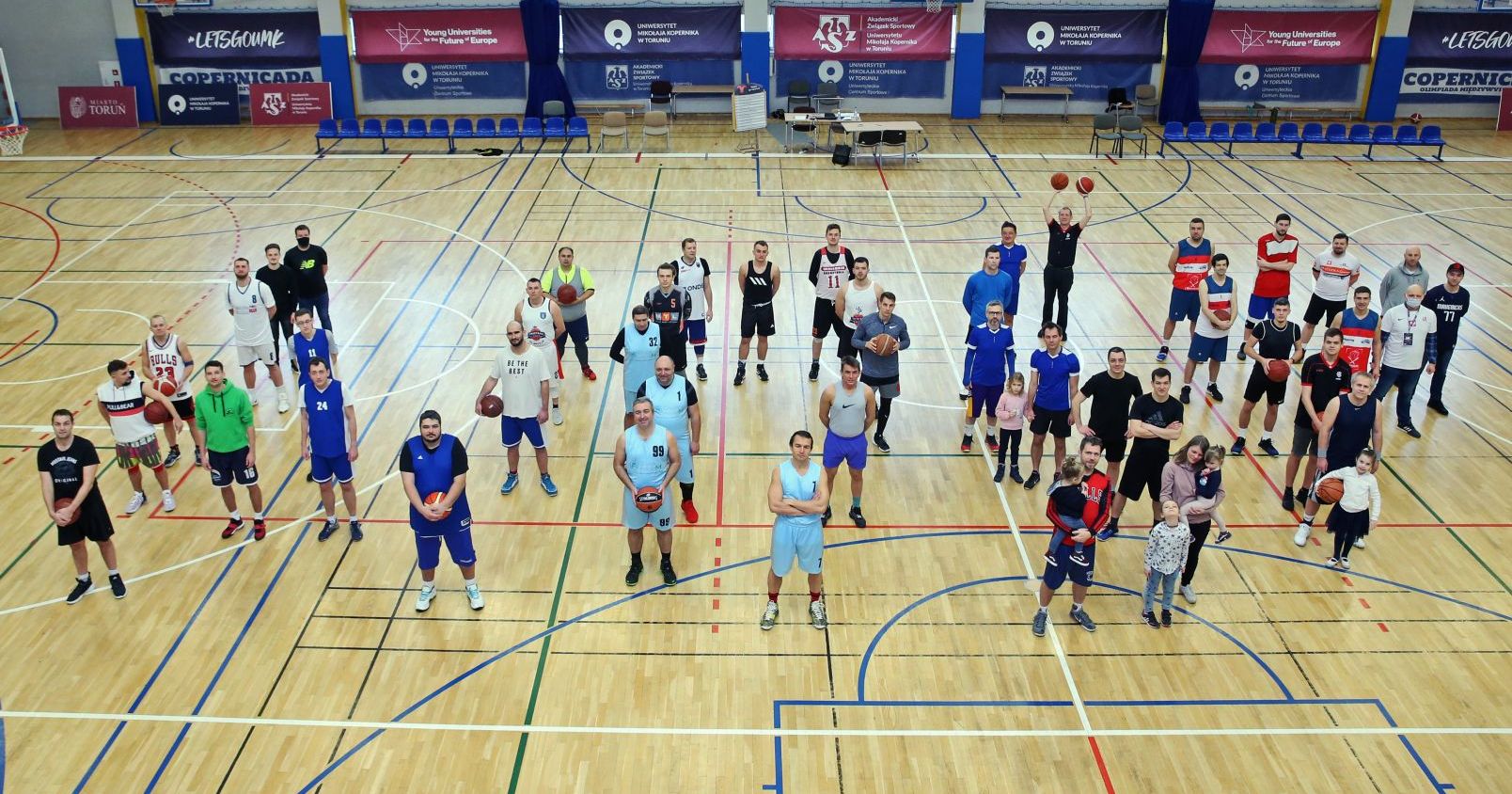 Uczestnicy turnieju koszykówki 3x3 w Uniwersyteckim Centrum Sportowym Grupa osób, zawodników koszykówki i kibiców stoi na parkiecie w hali sportowej