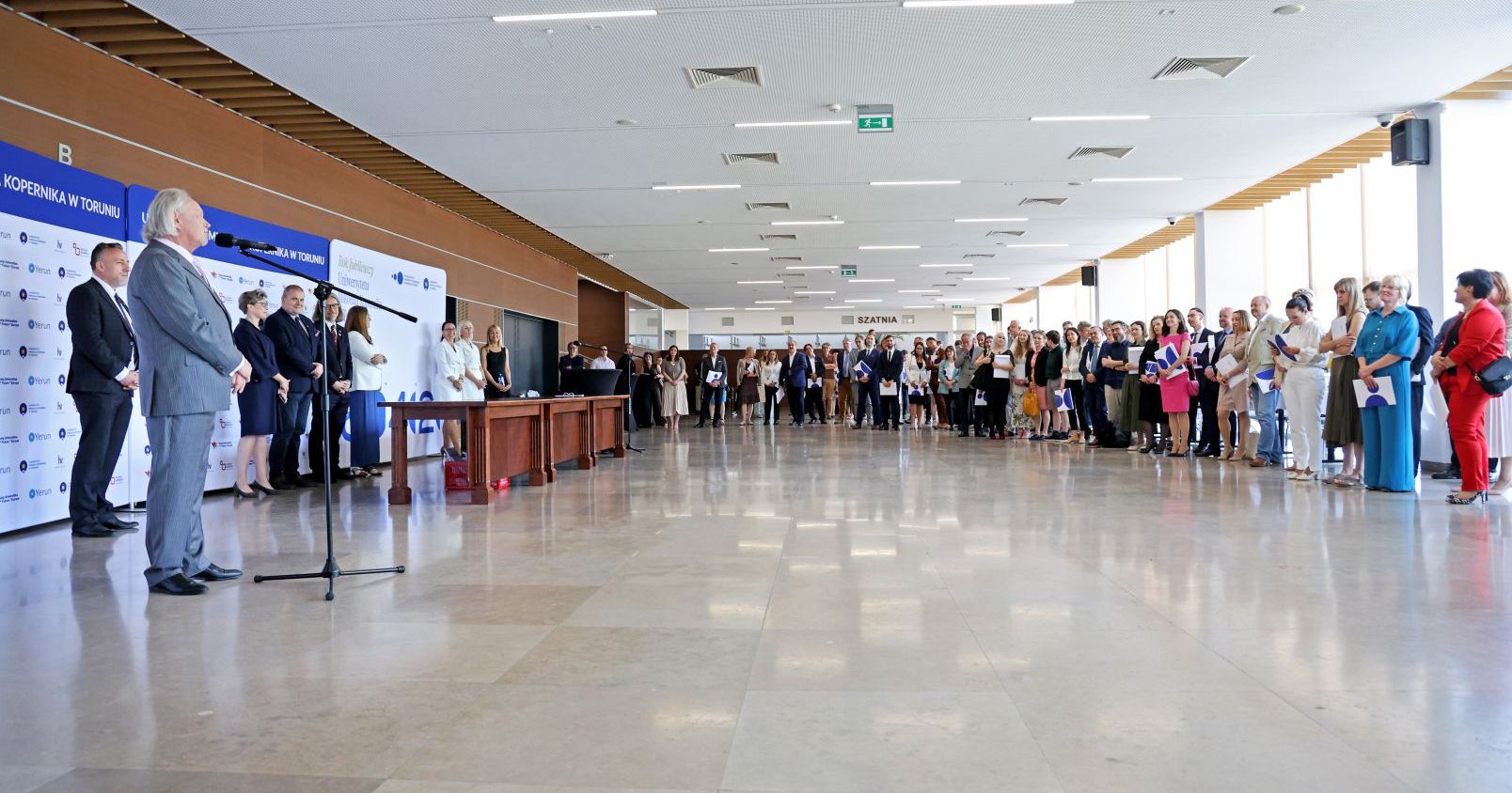Foyer Auli, rektor stoi przy mikrofonie, wokół duża liczba pracowników odbierających nagrody