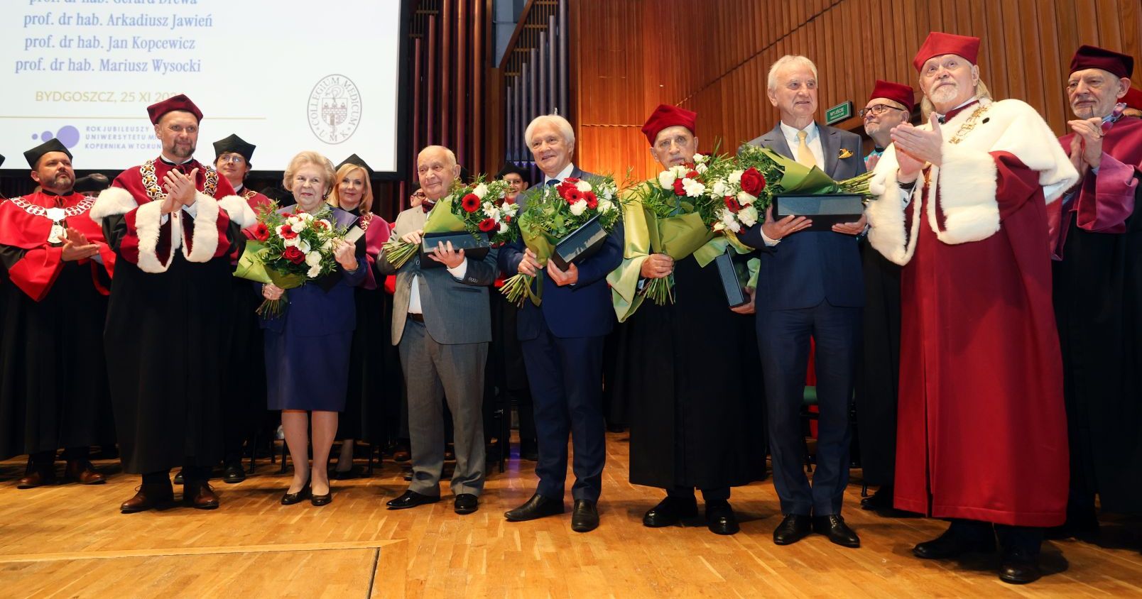 The first laureates of the Laurus Aesculapii award were Prof. Mieczysława Czerwionka-Szaflarska, Prof. Gerard Drewa, Prof. Arkadiusz Jawień, Prof. Jan Kopcewicz and Prof. Mariusz Wysocki Laureates of the Laurus Aesculapii award on stage along with prof. Andrzej Tretyn, NCU Rector and prof. Dariusz Grzanka, Vice-Rector for Collegium Medicum. Laureates are holding bouquets of flower and awards.