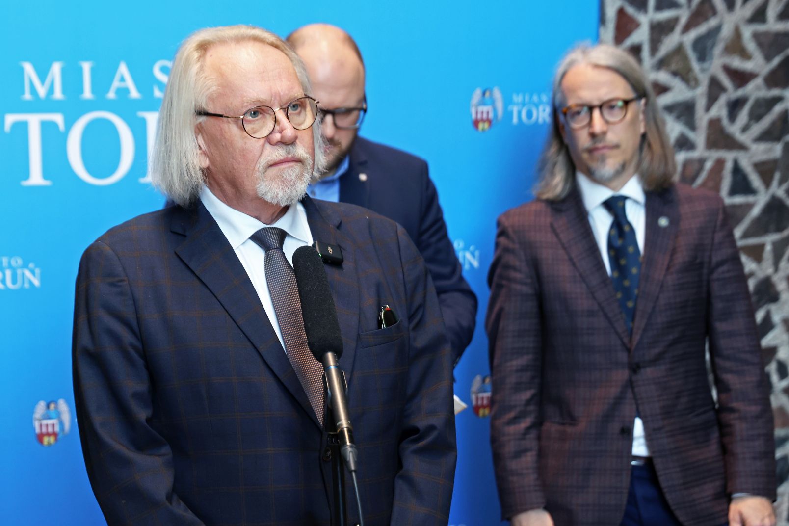 Prof. dr hab. Andrzej Tretyn, rektor UMK, stoi przy mikrofonie podczas konferencji prasowej. Za nim Paweł Gulewski, prezydent Torunia i dr hab. Adam Kola, prof. UMK, prorektor ds. nauki.