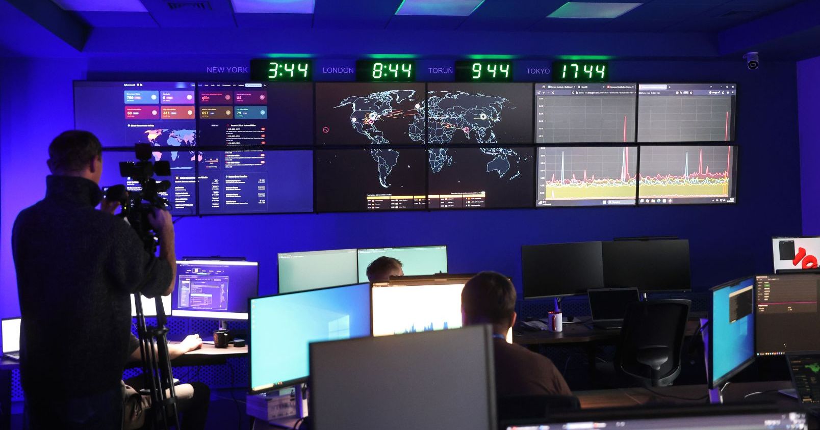 Nabór do Exea Cyber Akademii została zaprojektowana w modelu "skills first", czyli takiego, w którym liczą się realne umiejętności, a nie wpis w CV. Zegary pokazujące czas na świecie, pod nimi wiszą monitory i pracownicy siedzą przy biurkach z monitorami