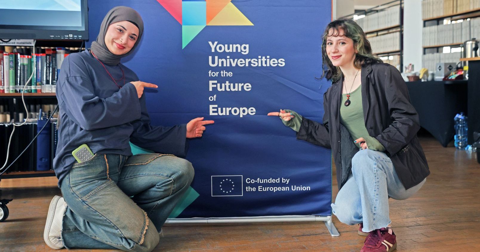 Osoby studiujące odgrywają centralną rolę we wszystkich etapach projektu YUFE. Gwarancją ich aktywnego udziału w rozwoju konsorcjum jest Studenckie Forum YUFE zrzeszające osoby reprezentujące wszystkie uczelnie partnerskie Dwie uśmiechnięte studentki przykucają i wskazują na logo YUFE na roll-upie