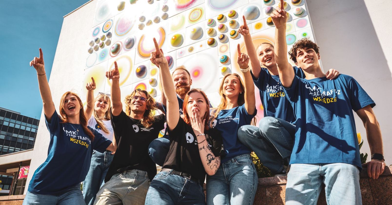 Each year, more and more people from abroad decide to study at the Nicolaus Copernicus University. Currently, there are over 1100 international students at the Nicolaus Copernicus University A group of male and female students wearing T-shirts with the NCU logo, reach up and point their finger at something. In the background, a characteristic mosaic on the façade of the NCU auditorium