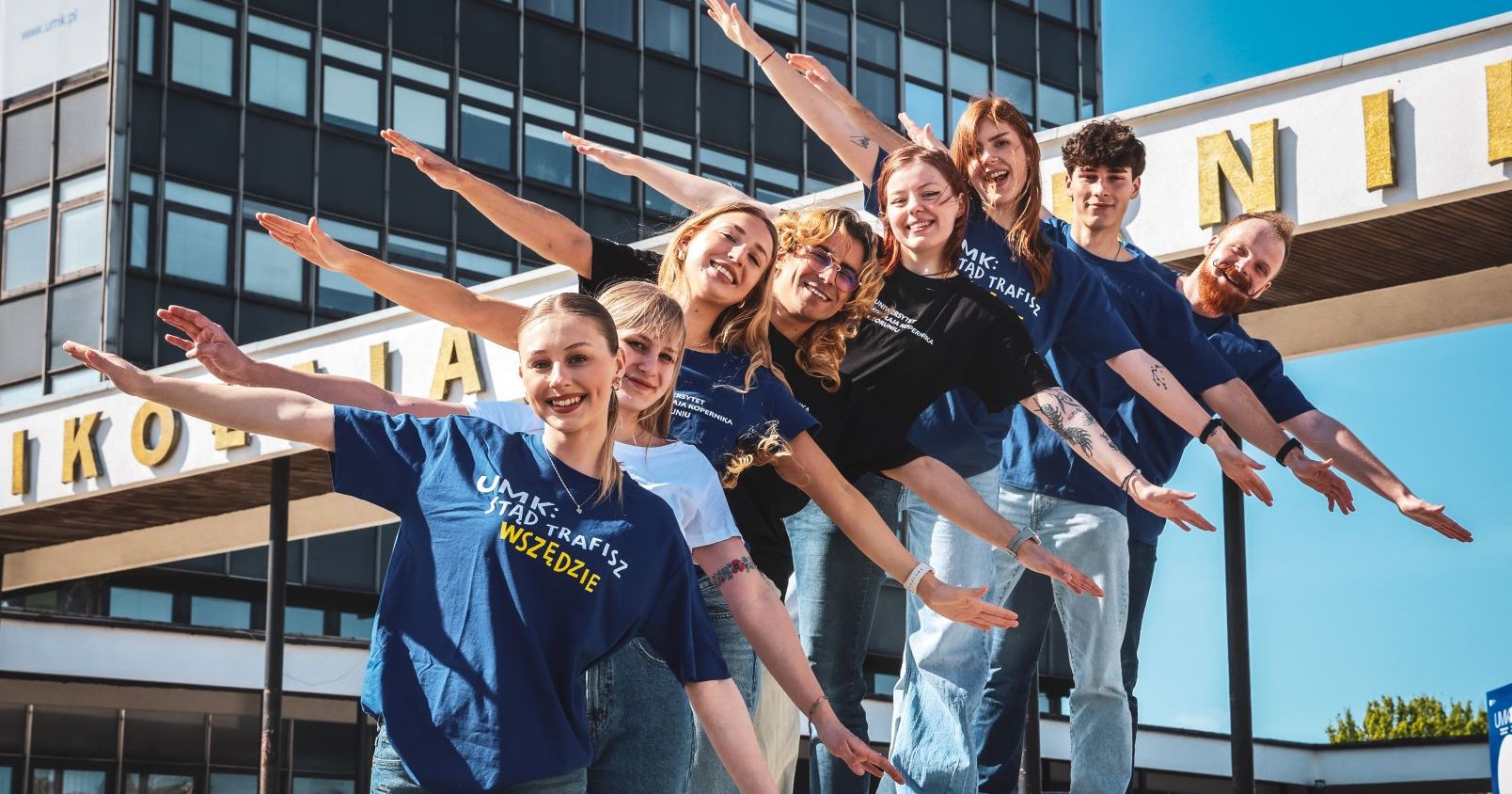 Przyszli studenci i studentki mogą wybierać z oferty ponad stu kierunków, w tym również studiów w języku angielskim oraz nowości Studenci z rozłożonymi ramionami, jakby szykowali się do lotu. W tle budynek rektoratu UMK