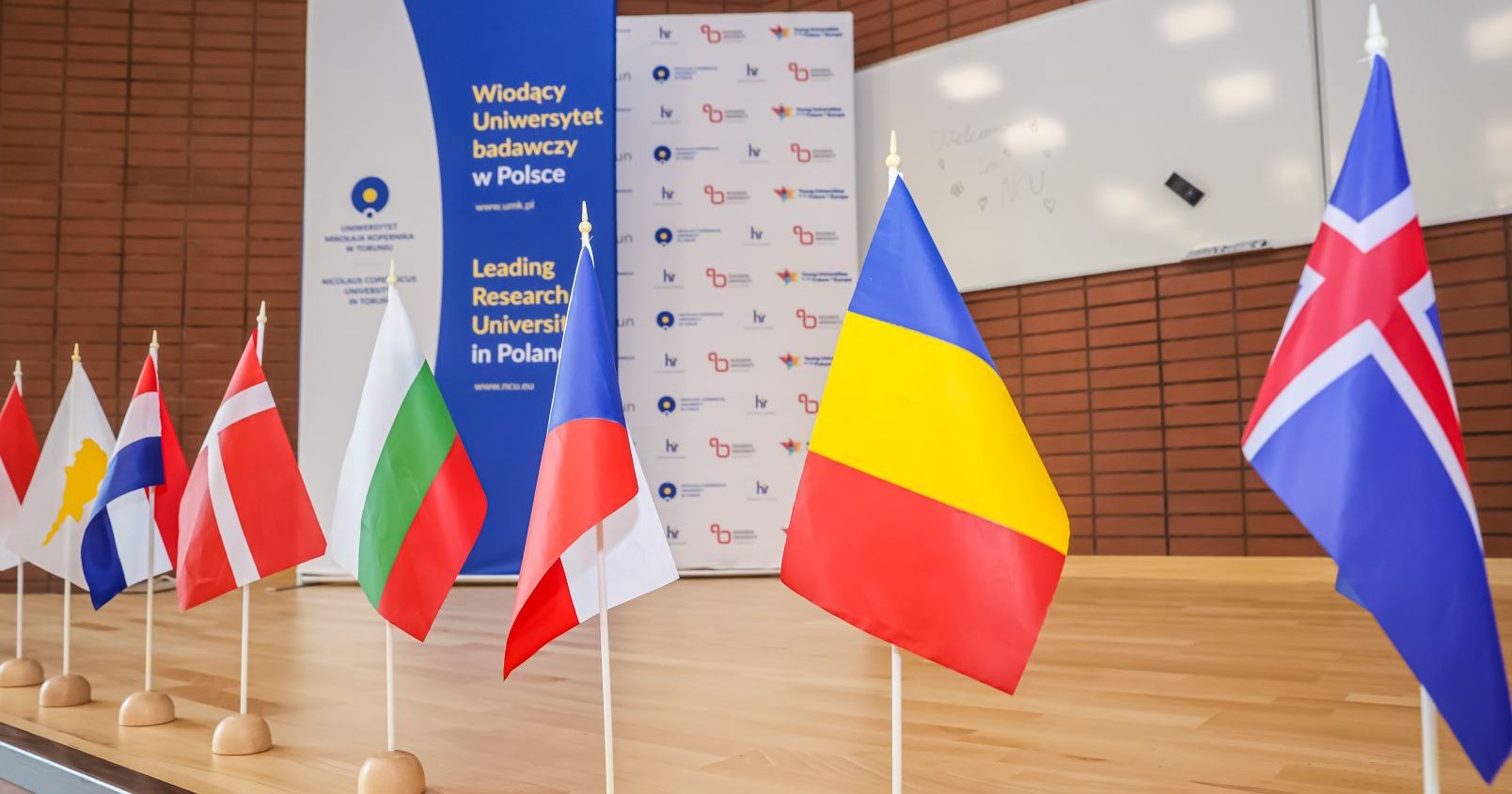 Małe flagi różnych krajów, a w tle rollup z logo UMK i napisem "wiodący uniwersytet badawczy w Polsce"