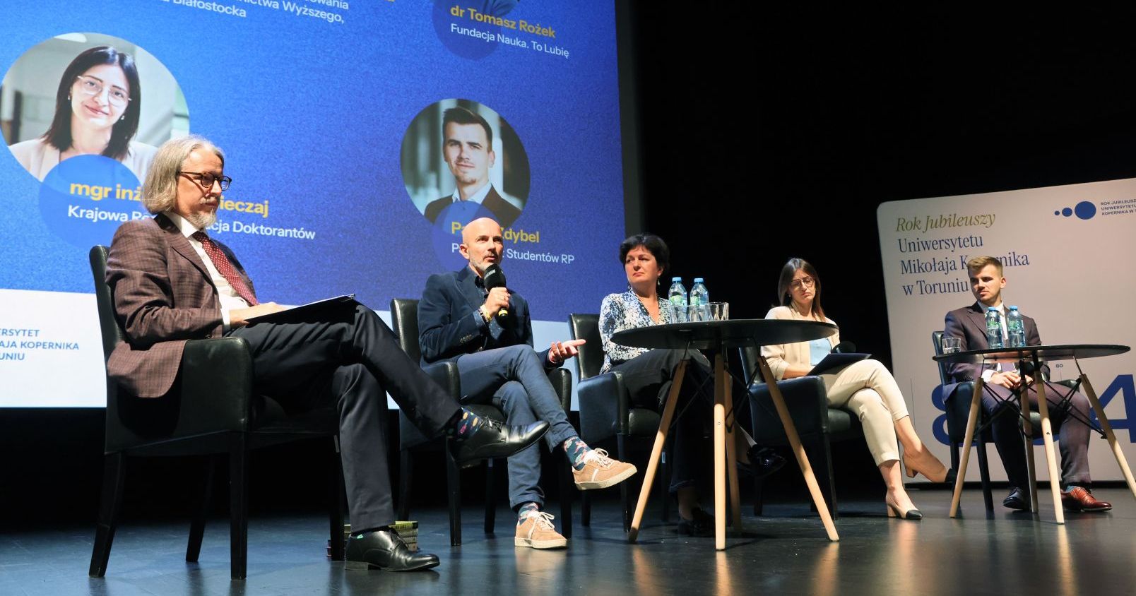 A group of people during a debate at Od Nowa, Dr Tomasz Rożek holding a microphone, in the background a wall with the inscription ‘Year of Jubilees of the Nicolaus Copernicus University in Toruń’