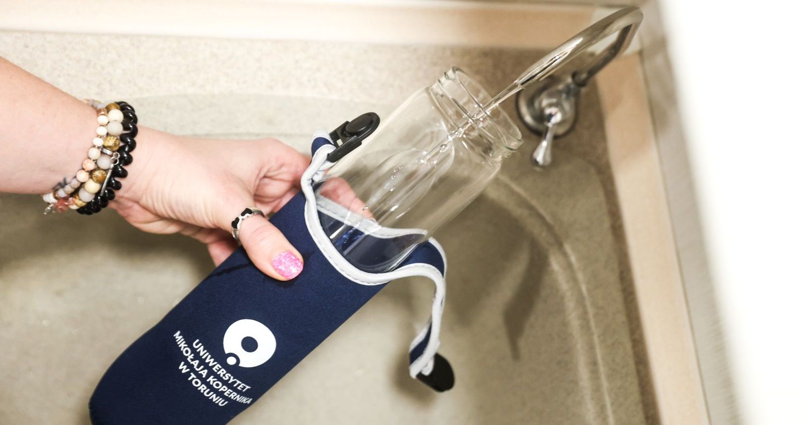 Ręka z butelką z logo UMK nalewająca wodę z nowego kranu do poboru wody pitnej