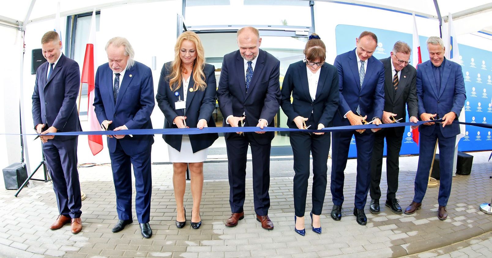 dwie kobiety i sześciu mężczyzn odświętnie ubranych przecina niebieską wstęgę przed drzwiami do budynku