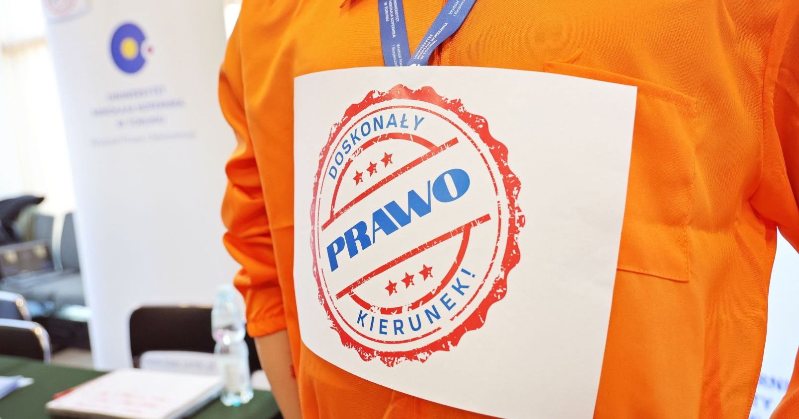 Grafika z napisem "Prawo doskonały kierunek" na kartce znajdującej się na pomarańczowym tle. Na drugim planie, który jest rozmyty, roll-up z logo Wydziału Prawa i Administracji