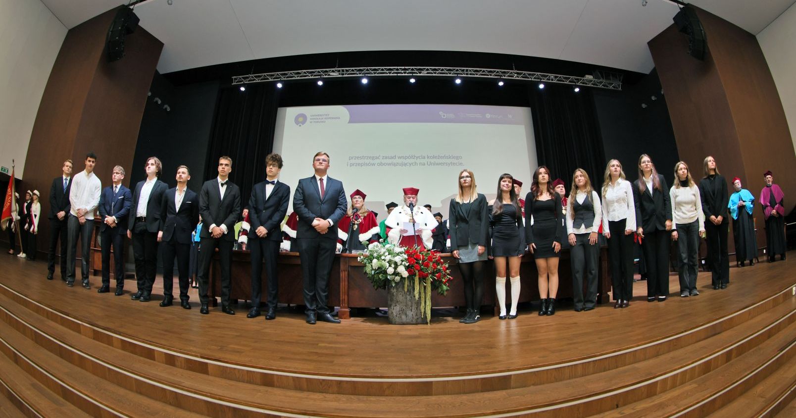 Szczególnie uroczystym punktem inauguracji była immatrykulacja, czyli symboliczne włączenie do społeczności akademickiej studentów i studentek I roku Immatrykulacja - nowo przyjęci studenci (kilkanascie osób) stoi w rzędzie na scenie Auli. Za nimi rektor odczytujący treść ślubowania
