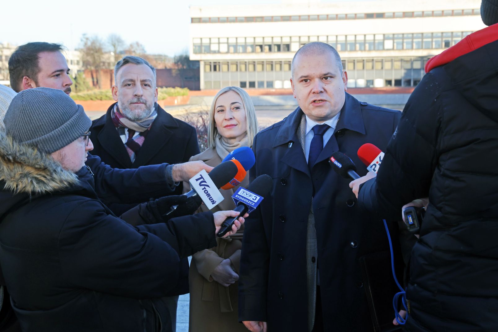 An outdoor scene of a group of reporters interviewing NCU Vice-Rector dr habil. Radosław Sojak. Standing behind him are mgr Karolina Sokalska and dr Patryk Tomaszewski. In the background you can see the NCU University Library.