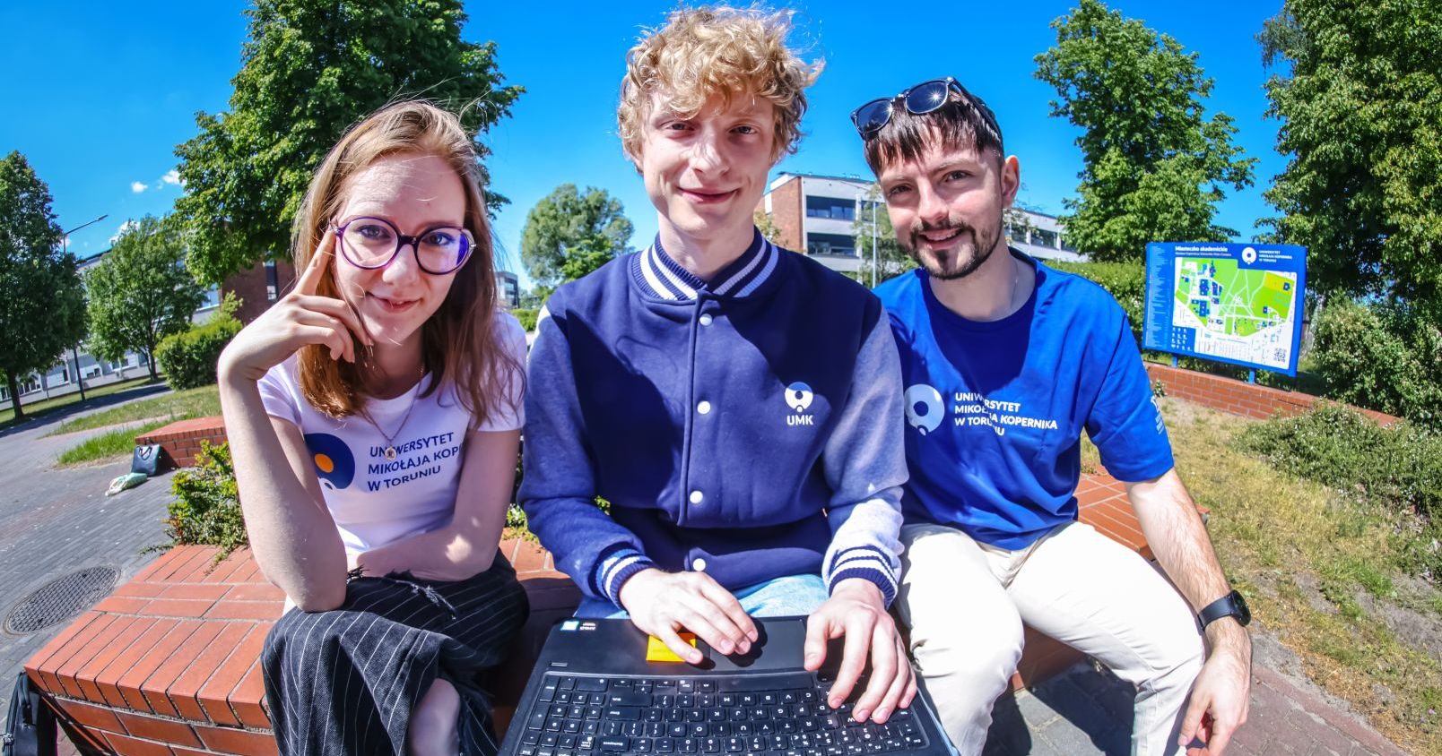 A female student and two male students are sitting next to each other, outside. The student sitting in the middle holds a laptop on his lap. The students are wearing clothes with the UMK logo