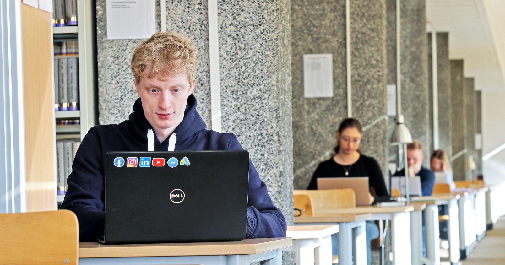 Na podium uczelni pod względem liczby stypendystów oprócz Uniwersytetu Mikołaja Kopernika w Toruniu stanęły Uniwersytet Warszawski (49) i Uniwersytet Jagielloński (37). Studenci i studentki w bibliotece siedzą przy biurkach z laptopami, po lewej stronie widać również fragment regału z książkami