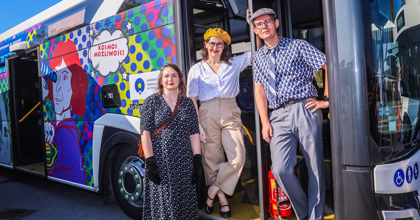  Colorfully wrapped bus with people standing by the door under bright sunlight.