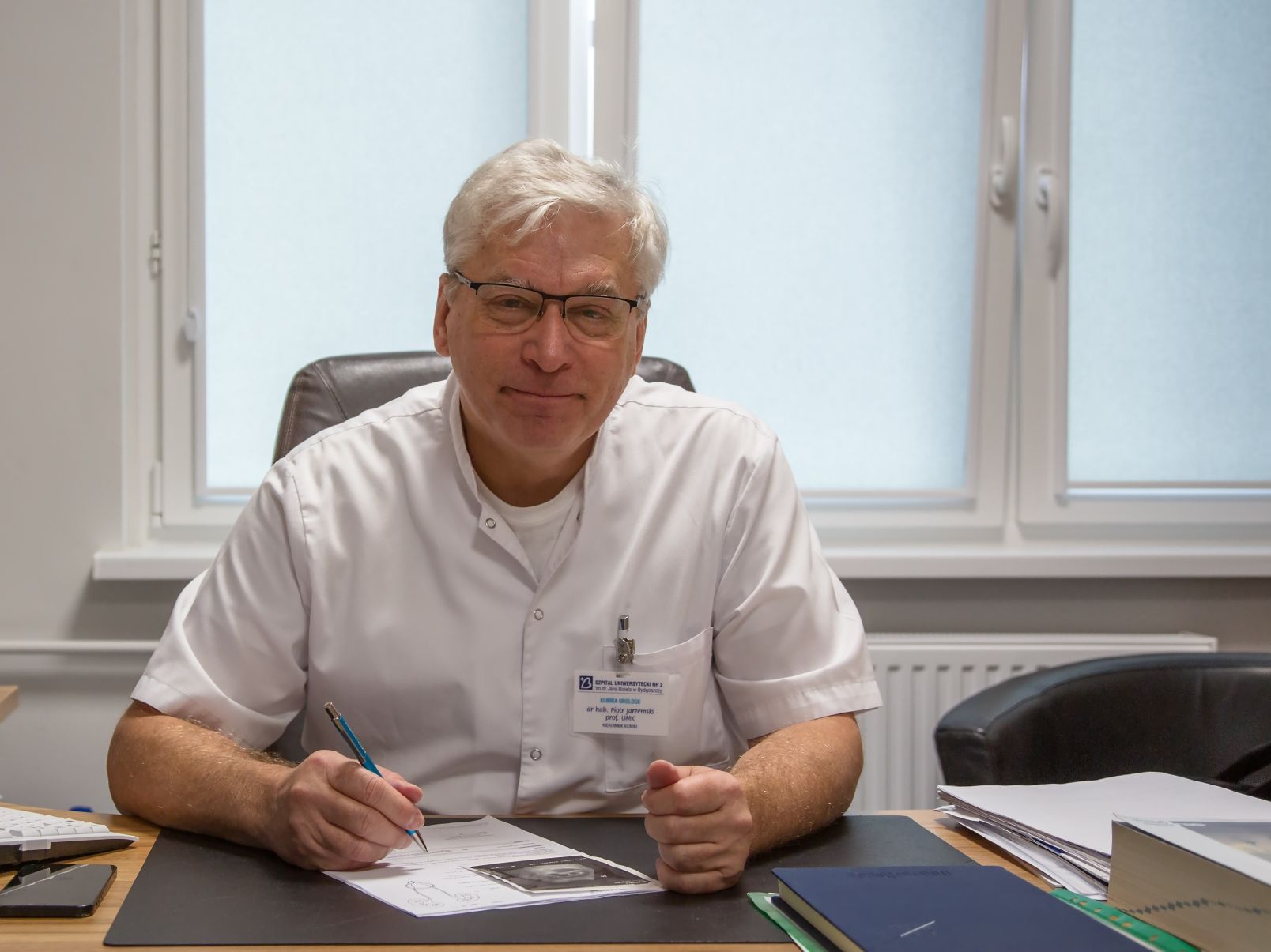 Dr hab. Piotr Jarzemski, prof. UMK z Katedry Urologii na Wydziale Nauk o Zdrowiu Collegium Medicum UMK siedzi w białym fartuchu przy biurku z długopisem w ręku