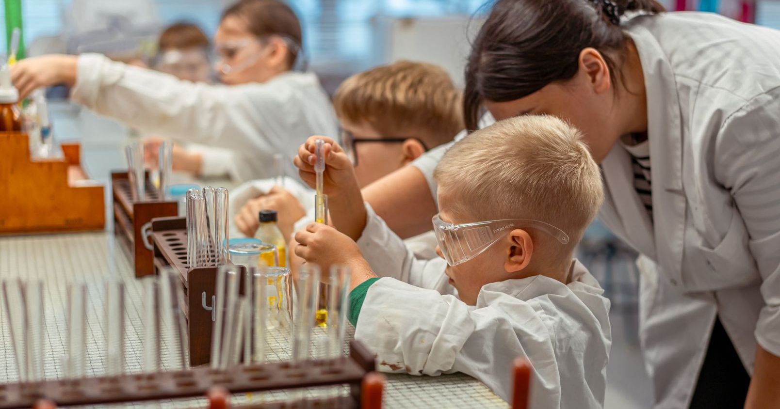 Organizatorami Toruńskiej Nocy Naukowców są Uniwersytet Mikołaja Kopernika w Toruniu i Centrum Nowoczesności Młyn Wiedzy Children in lab coats engaged in a science experiment with test tubes and beakers.