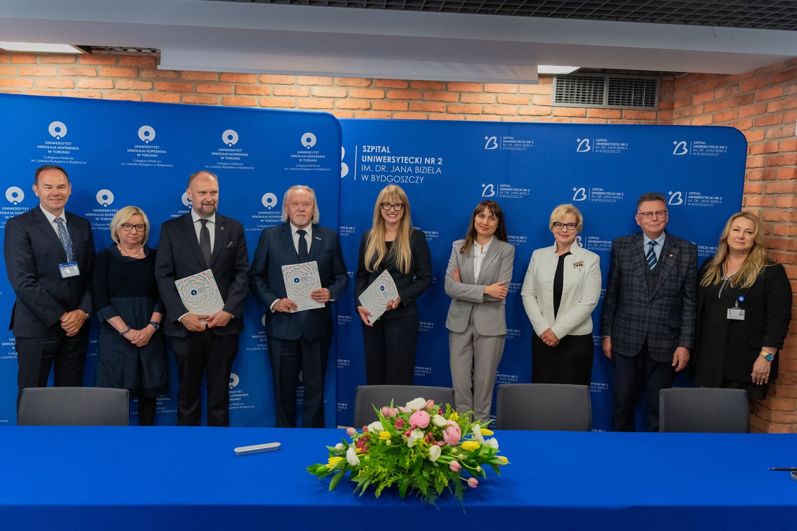Oficjele na tle niebieskiej ścianki UMK i szpitala Biziela podczas podpisania umowy na budowę Centrum Stomatologii UMK