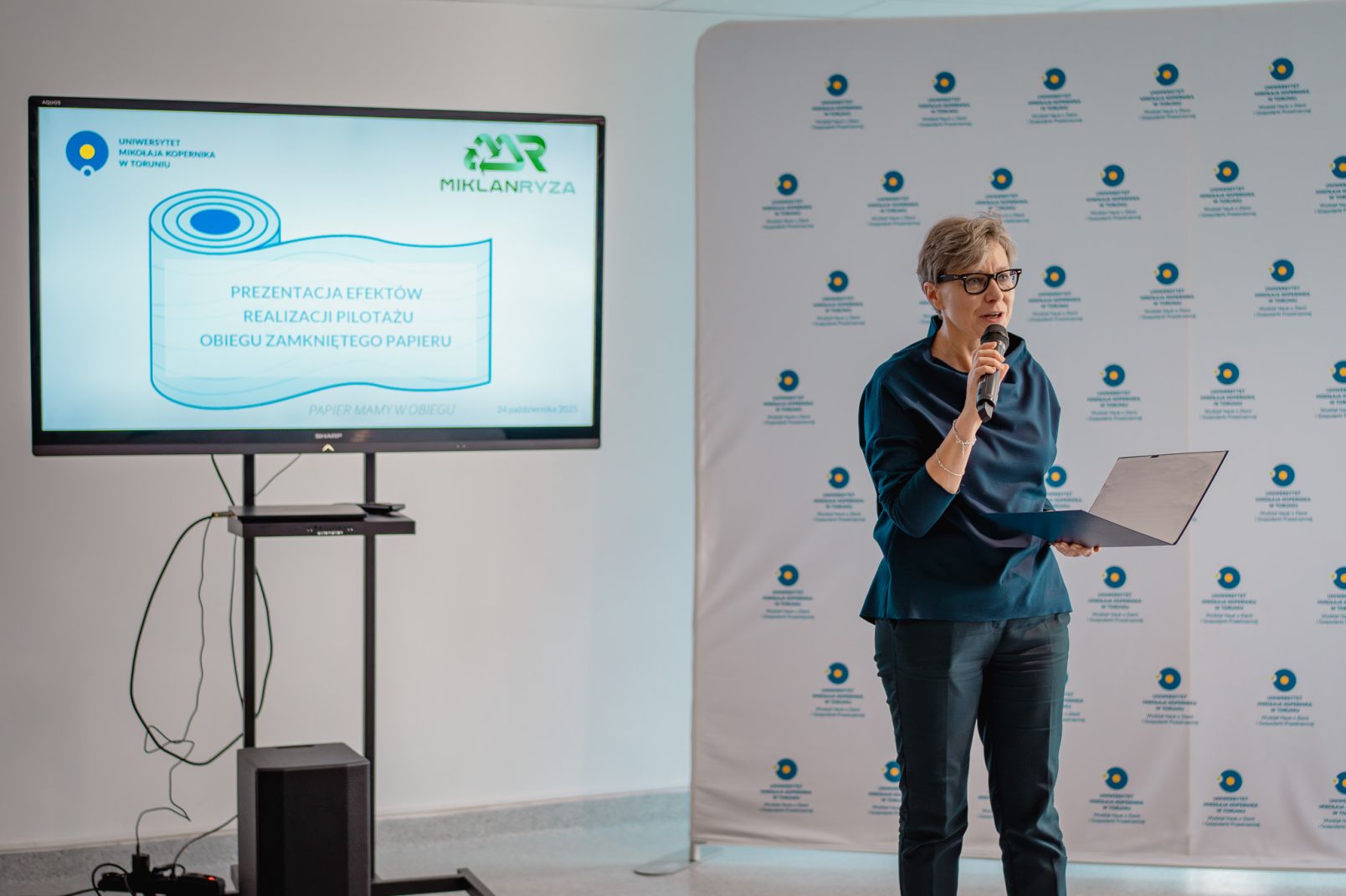 Prorektorka dr hab. Joanna Kucharzewska, prof. UMK stoi z mikrofonem na tle ścianki uniwersyteckiej i dużego telewizora z prezentacją