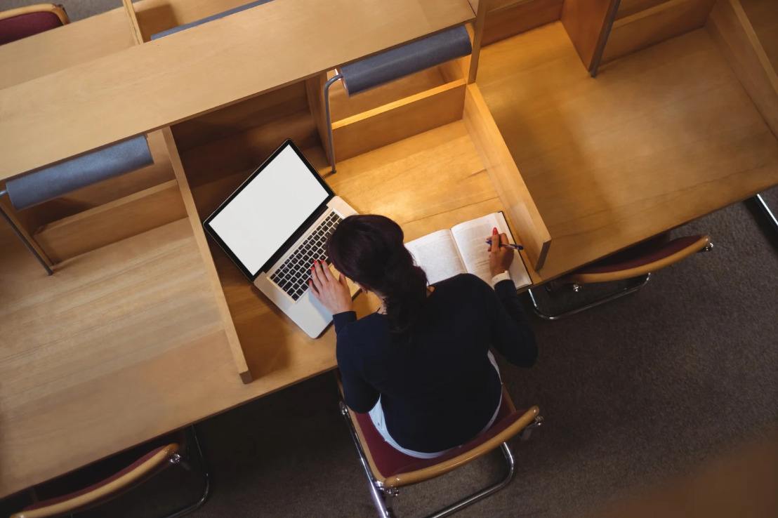 Pani siedzi w bibliotece, przed sobą ma notes, w którym coś zapisuje, i laptopa. Ujęcie z góry
