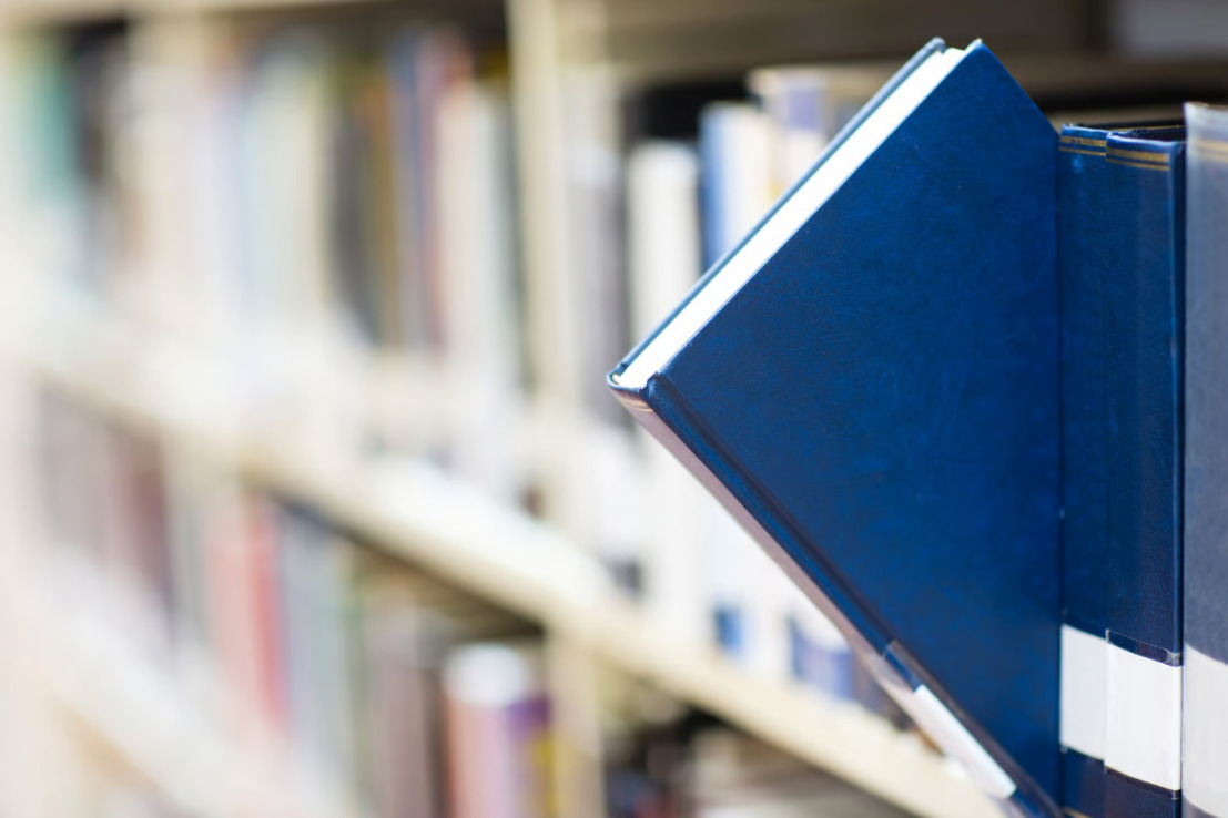 Jedna książka w gładkiej niebieskiej okładce wysunięta z rzędu innych książek na regale bibliotecznym