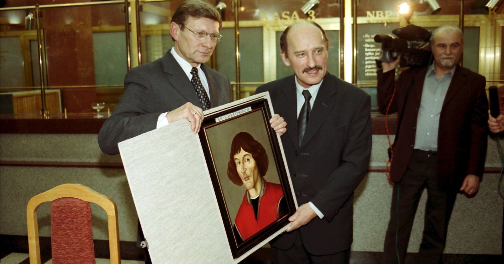 Leszek Balcerowicz i ówczesny rektor UMK Jan Kopcewicz trzymają portret Mikołaja Kopernika, stojąc w korytarzu NBP w Toruniu. W tle reporter z kamerą