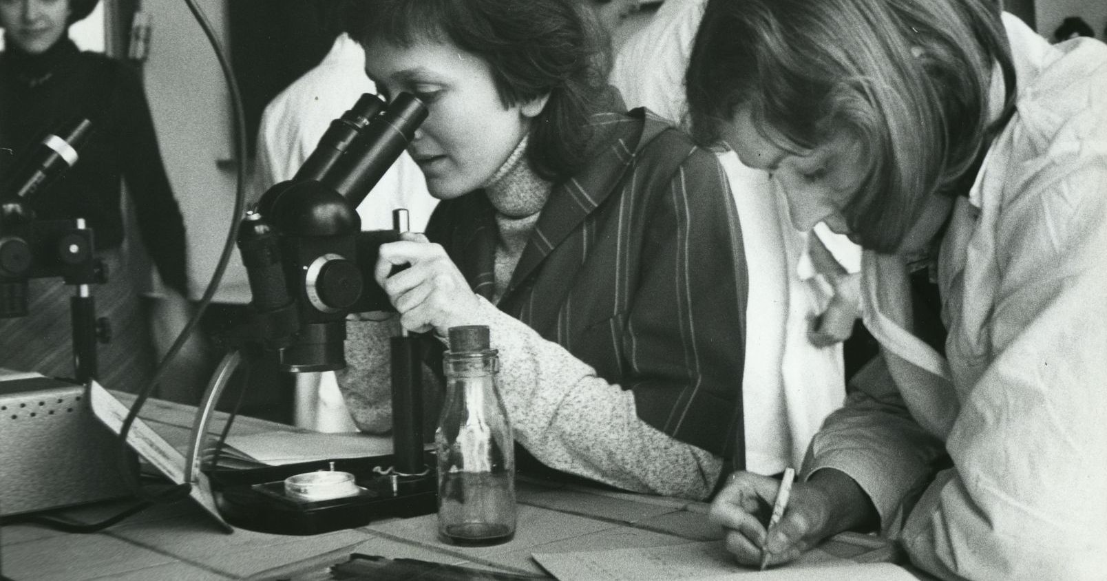 Stara fotografia w czerni i bieli przedstawiająca dwie studentki w laboratorium. Jedna z nich patrzy w mikroskop, druga robi notatki.