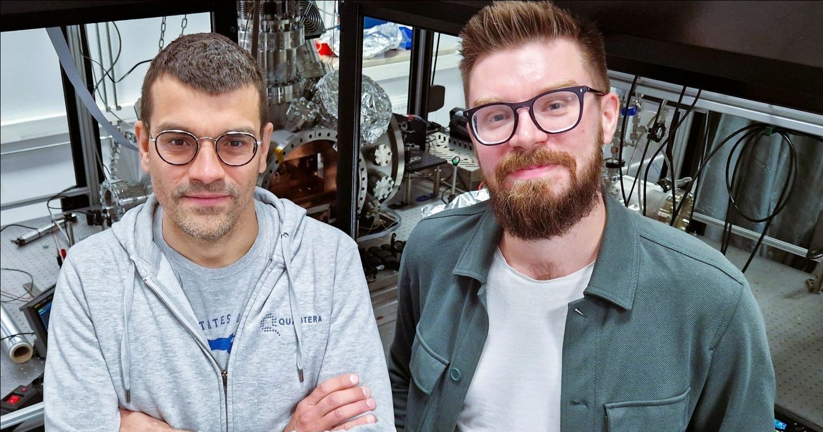 Prof. Piotr Wcisło and Dr. Hubert Jóźwiak stand side by side in their laboratory, with cables and specialized equipment visible in the background.
