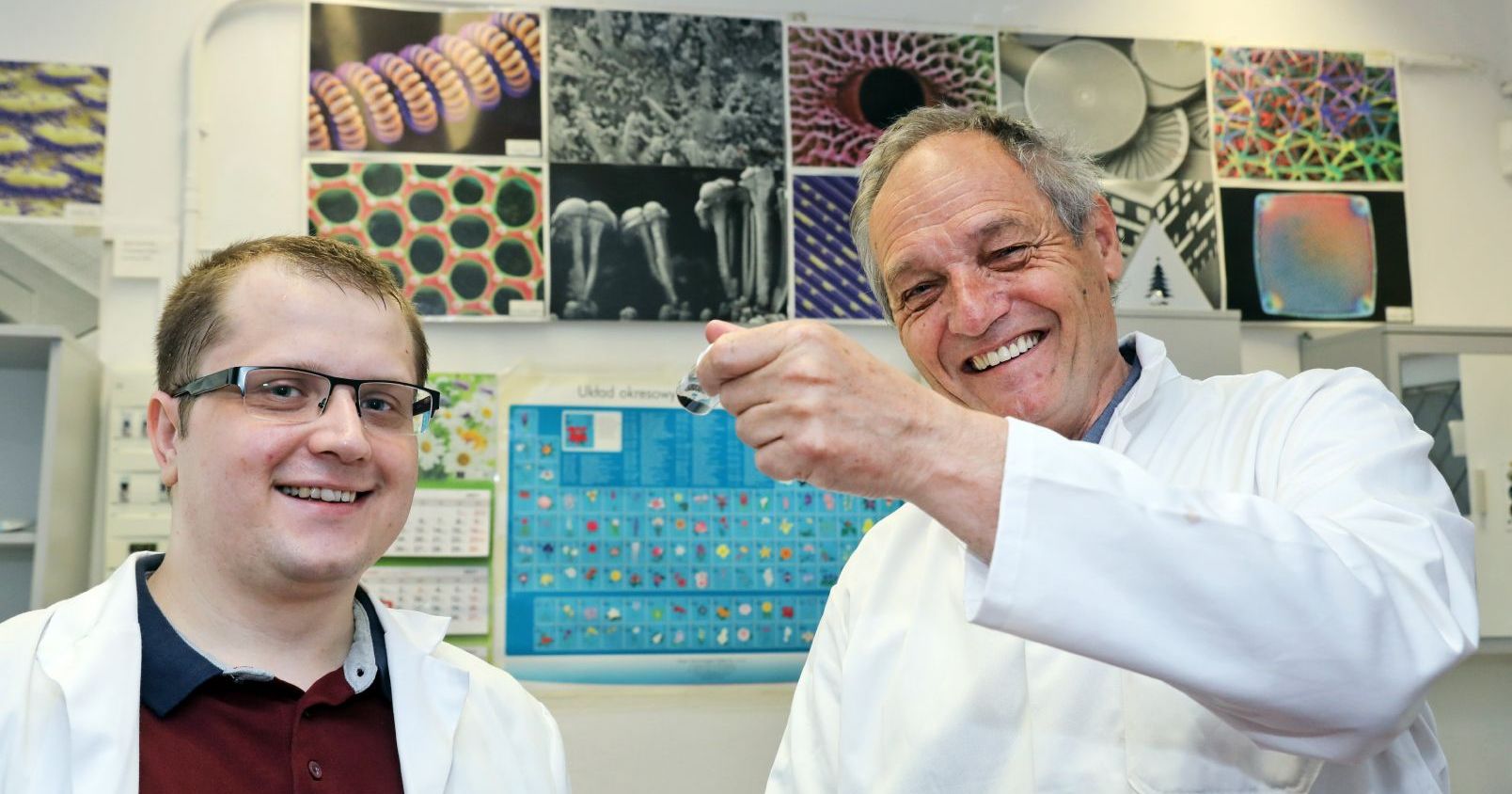 Dr Piotr Kamedulski i prof. dr hab. Jerzy P. Łukaszewicz Zdjęcie przedstawia dwóch uśmiechniętych mężczyzn. Mężczyzna po lewej stronie ma okulary. W tle wiszą zdjęcia różnych substancji widzianych przez mikroskop.
