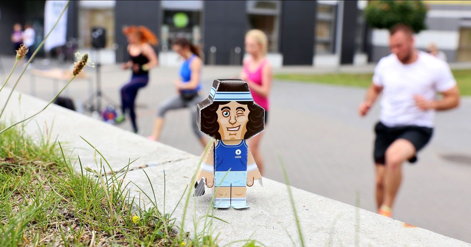 A folded paper figure of Copernicus as an athlete stands on a low wall. In the background, blurred people are engaged in sports