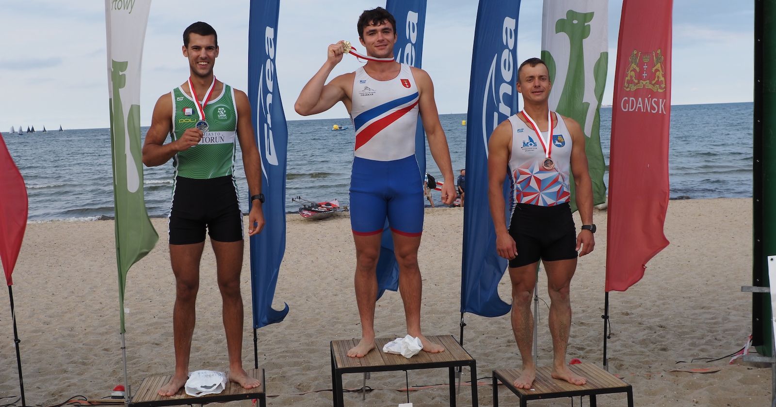 Razem z Kacprem Polańskim (po lewej) na podium stanęli także Adam Bujnowski (złoto) i Łukasz Zub (brąz) Trzech mężczyzn stoi na podium ustawionym na plaży. Za nimi stoją żagle reklamowe, a w tle widać także morze