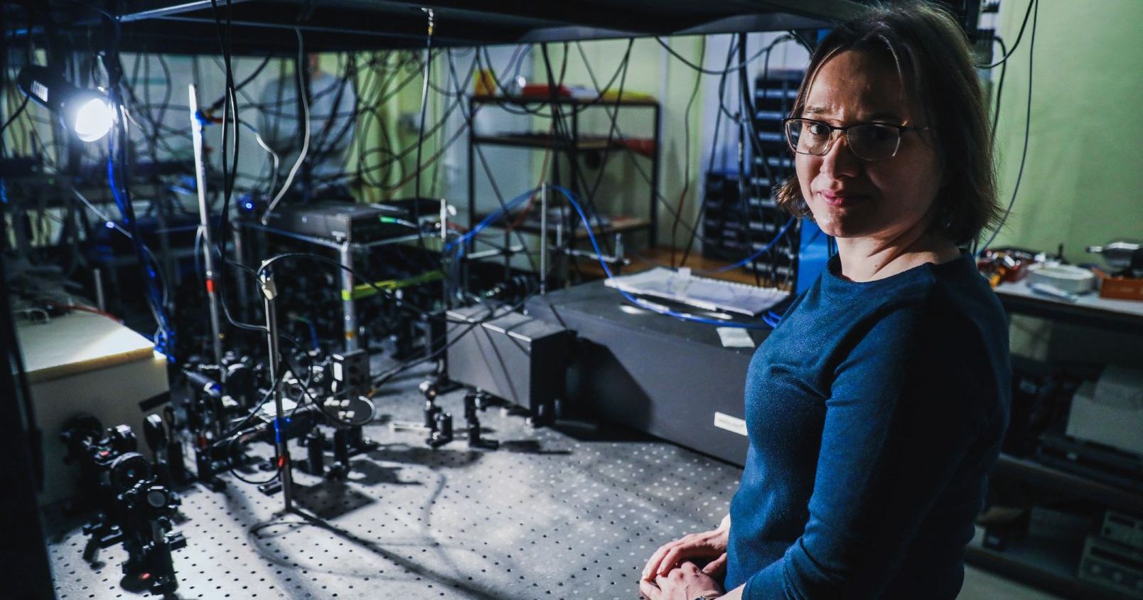 W laboratorium, pełnym sprzętów i kabli, po prawej stronie stoi kobieta w okularach