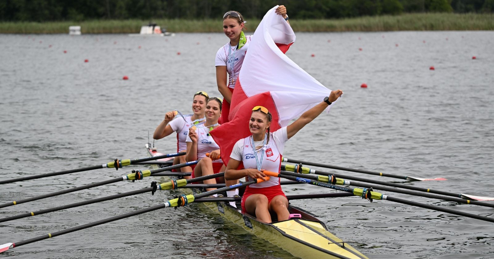 Osada reprezentantek Polski przekroczyła metę jako pierwsza, wyprzedzając reprezentację Czech i Wielkiej Brytanii Zawodniczki siedzą w łodzi po zwycięskim biegu, na szyjach mają medale, trzymają także polską flagę, jedna zawodniczka stoi