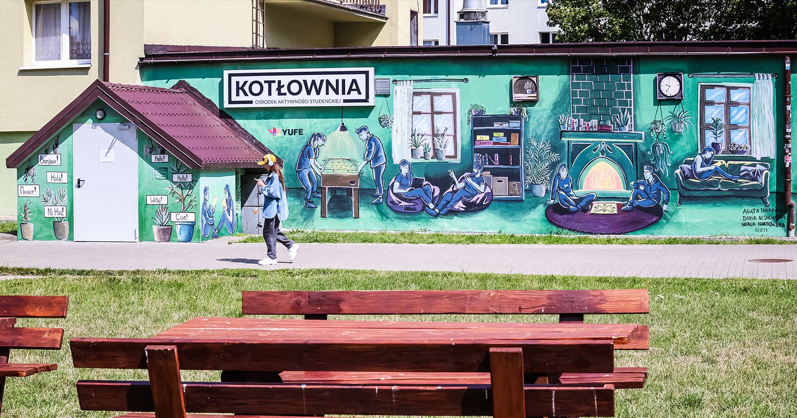 The new mural was created on the wall of the Student Activity Centre Kotłownia, which is located at Student House No. 5, 5/7 Słowackiego St. A mural on the facade of the Kotłownia, a person walks past it. There's also benches and a table in the foreground