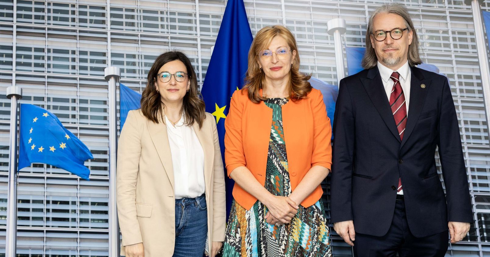 Stoją trzy osoby (dwie kobiety i jeden mężczyzna). W tle falgi Unii Europejskiej
