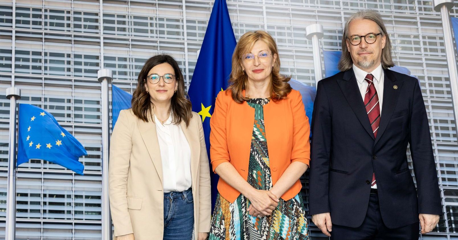 Three people stand (two women and one man). In the background, waves of the European Union
