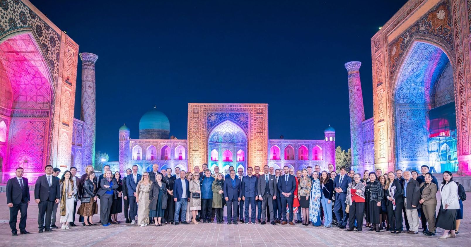Kilkadziesiąt osób stoi na tle charakterystycznej architektury Samarkandy. Jest wieczór, budynki są podświetlone różowo-niebiesko-fioletową iluminacją