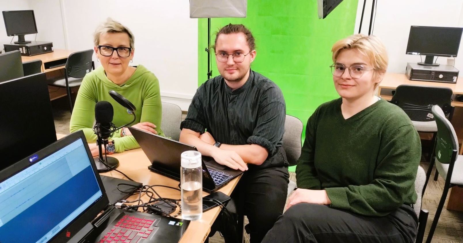 Dr hab. Magdalena Barwiołek, prof. UMK w towarzystwie zespołu redakcyjnego podcastu: Mateusza Szudry i Marii Lichograj Dr hab. Magdalena Barwiołek, prof. UMK w towarzystwie zespołu redakcyjnego podcastu: Mateusza Szudry i Marii Lichograj. Wszystkie osoby siędzą przy stole w sali komputerowej, na zdjęciu widać laptopy, mikrofon, statywy, a w tle greenbox