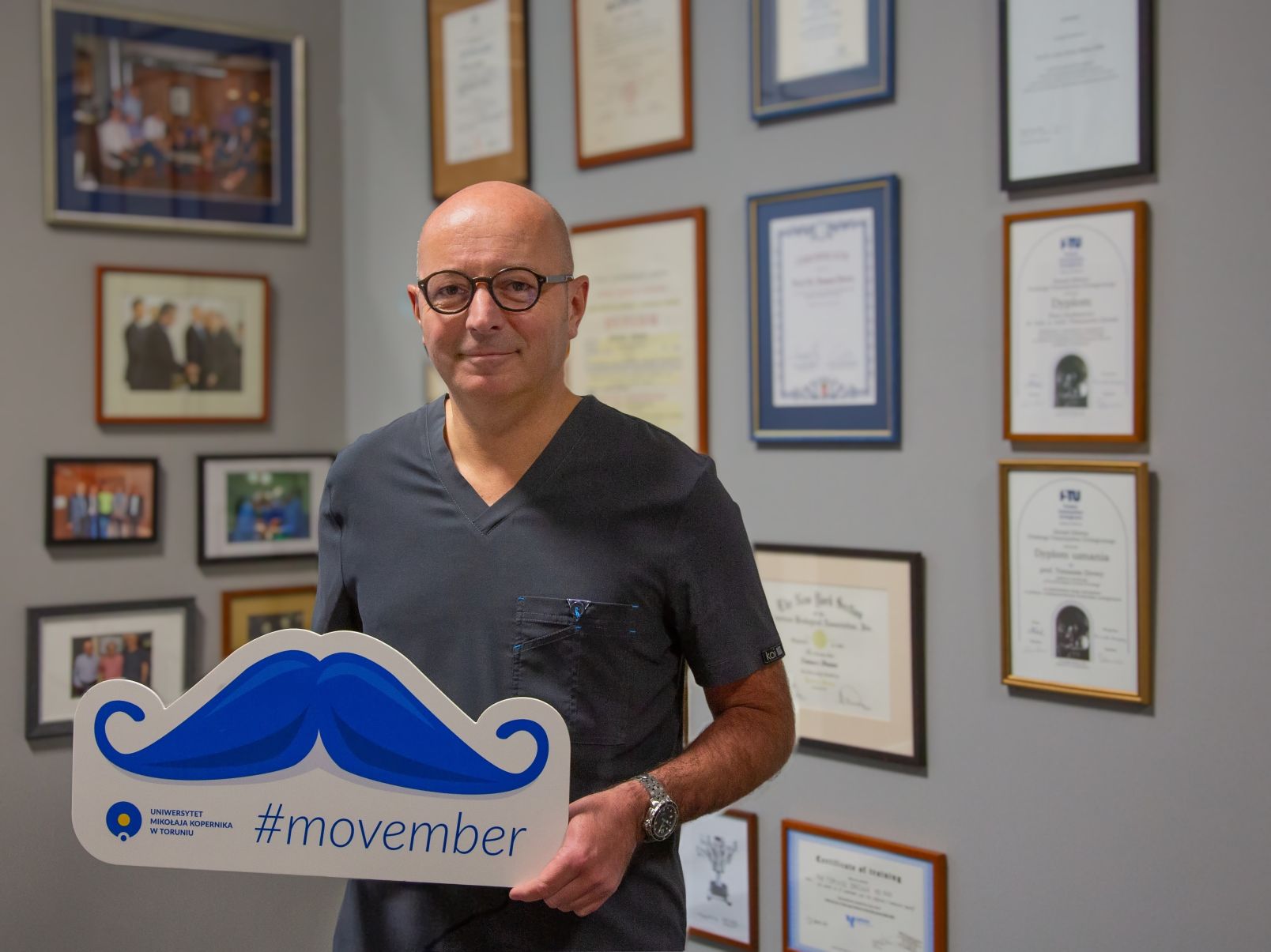 Prof. dr. hab. Tomasz Drewa, kierownik Katedry Urologii i Andrologii na Wydziale Lekarskim Collegium Medicum UMK stoi w gabinecie na tle zdjęć i dyplomów z tabliczką z wąsami i napisem movember